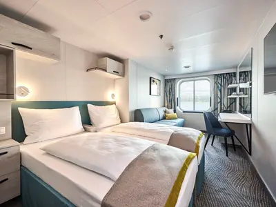 Innenansicht einer modernen Kabine der Mein Schiff 7 mit zwei einzeln stehenden Betten, einem Sofa am Fenster und einem Schreibtisch mit Stuhl. Eine große Fensterfront zeigt den Blick auf das Meer und eine bewaldete Küstenlinie. Die Kabine ist hell eingerichtet mit grauen und türkisen Akzenten.