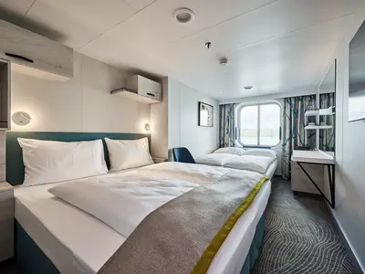 Ein breiter Blick auf eine moderne Kabine an Bord der Mein Schiff 7. Zwei Betten mit weißen Laken und einer grau-gelben Decke dominieren den Vordergrund. Ein rundes Fenster bietet eine Aussicht auf Wasser und Bäume. Links neben den Betten ist eine Kommode mit Leselampe und ein Wandschrank. Rechts befindet sich ein kleiner Schreibtisch mit Spiegel und ein großer Fernseher an der Wand.