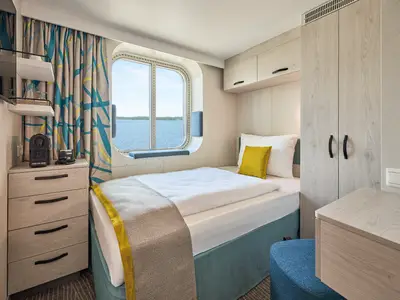 Innenansicht einer hellen und modernen Einzelkabine an Bord der Mein Schiff 7 mit einem Einzelbett und einem Fenster, das auf das Meer blickt. Links im Bild ist ein kleiner Nachttisch mit einer Kaffeemaschine zu sehen, rechts ein Kleiderschrank und eine kleine Kommode. Das Bett ist weiß bezogen und hat ein gelbes Kissen. Blaugraue Vorhänge mit gelben Akzenten hängen an der Wand.