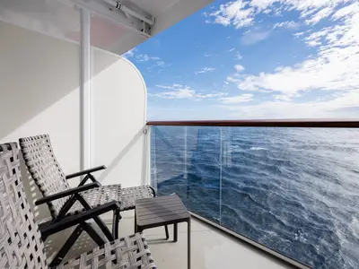 Sonniger Blick von einem Balkon der Mein Schiff 7 auf das tiefblaue, leicht bewegte Meer. Zwei graue Rattan-Sessel und ein kleiner Beistelltisch stehen auf dem Balkon. Die Reling aus Glas und Holz gibt den Blick frei auf den Horizont unter einem hellblauen Himmel mit vereinzelten weißen Wolken.