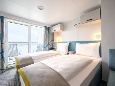 Innenansicht einer hellen Balkonkabine auf der Mein Schiff 7. Zwei getrennte Betten mit weißen Bezügen und grauen Überwürfen. Ein Balkon mit Stühlen bietet Blick auf das Wasser. Die Vorhänge sind blau-gelb gemustert. An der Wand befinden sich Schränke und Leselampen.