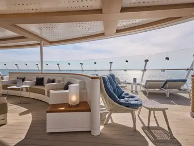 Blick auf das Außendeck der Mein Schiff 7 mit bequemen, gepolsterten Lounge-Möbeln und Sonnenliegen. Im Hintergrund ist das blaue Meer unter einem hellen Himmel zu sehen. Eine Glasbrüstung trennt das Deck vom Ozean.