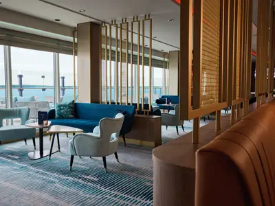 Blick in eine stilvolle Lounge der Mein Schiff 7 mit großen Fenstern zum Meer. Blaue Sofas und helle Sessel stehen auf einem gestreiften Teppich, durch goldene Trennelemente abgetrennt.