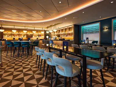 Innenansicht der Bar an Bord der Mein Schiff 7 mit geometrischem Fußboden, hohen Tischen und Stühlen in Grau und Dunkeltürkis. Eine lange Bar mit vielen Flaschen ist im Hintergrund beleuchtet, während an der Wand Fenster mit türkisfarbenen Rollos und goldene Wandleuchten zu sehen sind.