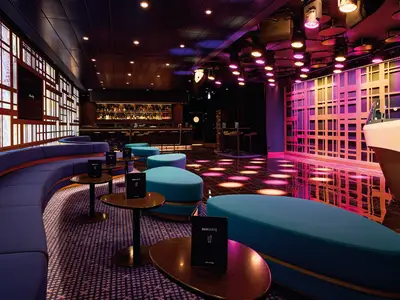 Innenansicht einer modernen Bar auf der Mein Schiff 7 mit lila Rundsofas, türkisfarbenen Hockern, hohen Tischen und einer Bar. Die Decke ist dunkel mit vielen Lichtern. Die Wände sind mit Leuchtstreifen und Mustern versehen. Der glänzende Boden reflektiert die farbige Beleuchtung.