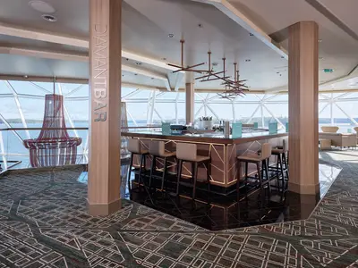 Ein Panoramabild der Diamant Bar an Bord der Mein Schiff 7. Die moderne Bar mit dunklem Holz und goldenen Akzenten ist von großen Fenstern umgeben, die einen weiten Blick auf das Meer und die Küste freigeben. Elegante Barhocker und eine gemütliche Sitzecke ergänzen das Ambiente.