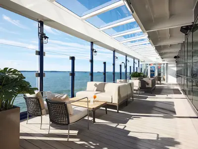 Blick auf das Außendeck der Mein Schiff 7: Eine helle Holzterrasse mit modernen Lounge-Möbeln, umgeben von einer Glasbrüstung. Ein großes Meer erstreckt sich bis zum Horizont unter einem blauen Himmel. Eine Pflanze steht im Vordergrund, und im Hintergrund sind weitere Sitzgelegenheiten und eine Glaswand sichtbar.