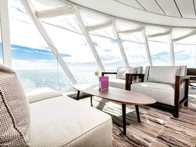Eine helle und moderne Lounge an Bord der Mein Schiff 7 mit großen Fenstern und weitem Blick auf das blaue Meer und den Himmel. Bequeme Sessel und Sofas mit hellgrauen Bezügen und ein Holztisch mit einer rosa Menükarte und einer Vase mit einer grünen Blume sind zu sehen. Die Holzvertäfelung und der Teppichboden in Naturtönen tragen zur entspannten Atmosphäre bei. Es fällt viel Sonnenlicht in den Raum.