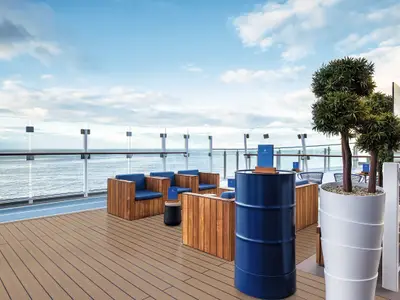 Blick von einer Lounge auf dem Oberdeck der Mein Schiff 7: Holzmöbel mit blauen Kissen stehen auf einem Holzboden. Dahinter erstreckt sich das weite, blaue Meer bis zum Horizont unter einem hellblauen Himmel mit einigen Wolken. Eine durchsichtige Glasbrüstung sichert das Deck zum Meer hin ab. Ein großer Topf mit zwei kleinen Bäumen steht rechts. In der Mitte des Bildes steht ein blauer Tisch in Form eines Fasses.