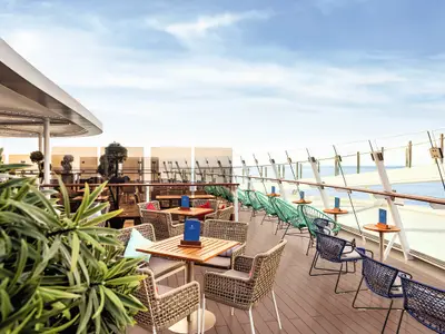 Sonniger Outdoor-Loungebereich an Deck der Mein Schiff 7 mit Holztischen, Rattanstühlen und modernen, türkisfarbenen und blauen Sesseln, umgeben von Pflanzen und einer Glasbrüstung mit Blick auf das blaue Meer.