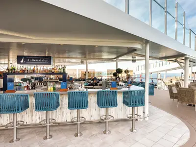 Blick auf die Aussenalster Bar an Bord der Mein Schiff 7, einer Outdoor-Bar mit blau-türkisen Barhockern und einer gut bestückten Bar. Im Hintergrund sind Loungesessel und das offene Meer zu sehen.