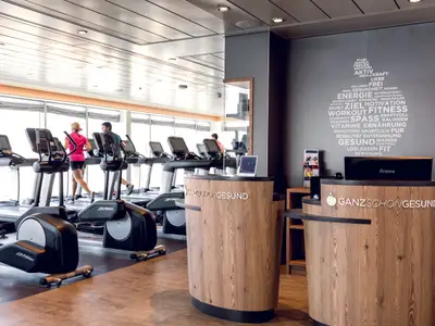 Blick in den modernen Fitnessraum der Mein Schiff 7. Im Vordergrund mehrere Laufbänder und Crosstrainer. Im Hintergrund trainieren zwei Personen auf Laufbändern vor großen Fenstern mit Meerblick. Rechts ist eine Rezeption aus hellem Holz mit dem Schriftzug "GANZSCHÖNGESUND" und einem Apfellogo zu sehen. An der Wand darüber befindet sich eine wortförmige Grafik zum Thema Gesundheit.