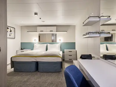 Blick in eine moderne Kabine der Mein Schiff 7: Im Zentrum steht ein Doppelbett mit weißer Bettwäsche und grauer Decke. Links ein kleiner Nachttisch mit Telefon, rechts ein weiterer mit Schubladen. Über dem Bett ein teal-farbenes Kopfteil und Spiegel. Rechts im Vordergrund ein Schreibtisch mit Stuhl und Kaffeemaschine.