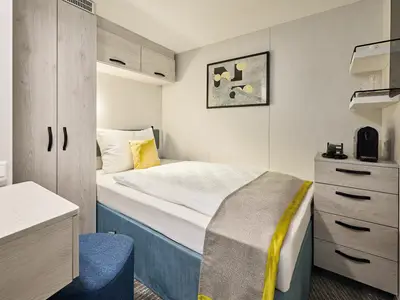 Ein Blick in eine modern eingerichtete Einzelkabine an Bord der Mein Schiff 7. Man sieht ein Bett mit weißer Bettwäsche und gelbem Kissen. Links stehen ein heller Schrank und ein kleiner Tisch. Rechts befindet sich ein Nachttisch mit Nespresso-Maschine unter schwebenden Regalen.