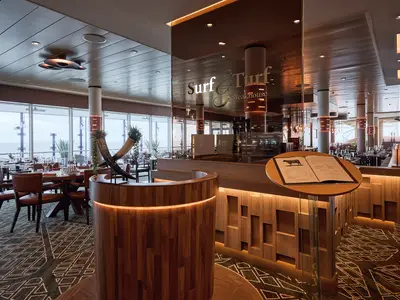 Innenansicht des Surf & Turf Steakhouse auf der Mein Schiff 7. Elegante Holzeinrichtung, Ledersessel und Tische sind zu sehen. Durch große Fenster im Hintergrund blickt man auf das offene Meer.