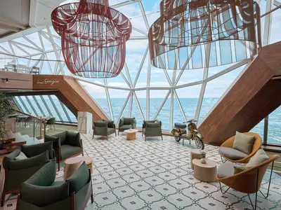 Blick in die moderne "La Spezia" Lounge auf der Mein Schiff 7 mit riesigen Panoramafenstern mit Meerblick. Grüne Sessel und beigefarbene Tische stehen auf einem gemusterten Boden. Eine goldene Vespa ist Teil der Dekoration. Im Hintergrund ist ein italienisches Restaurant zu sehen.