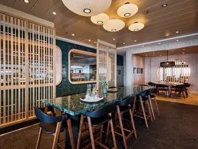Innenansicht eines modernen Restaurants auf der Mein Schiff 7: links offene Küche hinter Holzlamellen, mittig langer Marmortisch mit Barhockern, rechts Essbereich mit runden Tischen und einem runden Fenster.