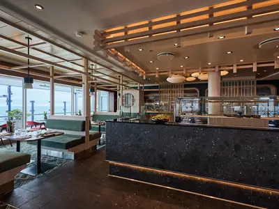 Innenansicht eines stilvollen Restaurants an Bord der Mein Schiff 7. Grüne Sitzbänke und Holztische sind am Fenster mit Meerblick aufgestellt. Im Vordergrund eine schwarze Theke mit Vitrine, darüber ein Deckenbereich mit Holzelementen und Hängelampen.
