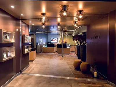 Blick in den luxuriösen SPA & MEER Bereich an Bord der Mein Schiff 7. Links sind beleuchtete Vitrinen mit Produkten zu sehen. Im Hintergrund führen verschiedene Holzböden zu einer Rezeption mit einem Bildschirm, der eine liegende Frau zeigt. Rechts stehen große Pflanzen und eine Wand mit der Aufschrift 'SPA & MEER' und 'Zum Saunabereich'. Eine Laterne mit Kerze und zwei Hocker befinden sich im Vordergrund.