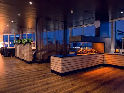 Ein stilvoller Innenbereich der Mein Schiff 7 in warmem Licht. Man sieht eine moderne Lounge mit Holzboden, einem beleuchteten Kamin mit Holzscheitimitation und Kerzen. Grüne Pflanzen in hohen Pflanzgefäßen und bequeme Sitzgelegenheiten sind zu sehen. Große Fenster geben den Blick auf das Meer frei. Die Uhrzeit ist 12 Uhr 10.