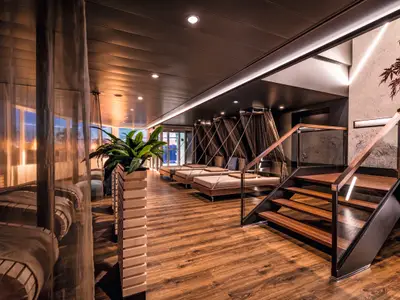Ein luxuriöser Entspannungsbereich an Bord der Mein Schiff 7 mit dunklem Holzfußboden. Rechts führt eine moderne Treppe mit Glasgeländer und Holzhandlauf nach oben. Links stehen mehrere Hängebetten mit braunen Vorhängen und bequemen Auflagen. Im Vordergrund befindet sich eine hohe Pflanze in einem Holzgefäß. Die Decke ist dunkel gehalten mit integrierter Beleuchtung. Fenster im Hintergrund lassen diffuses Licht herein.