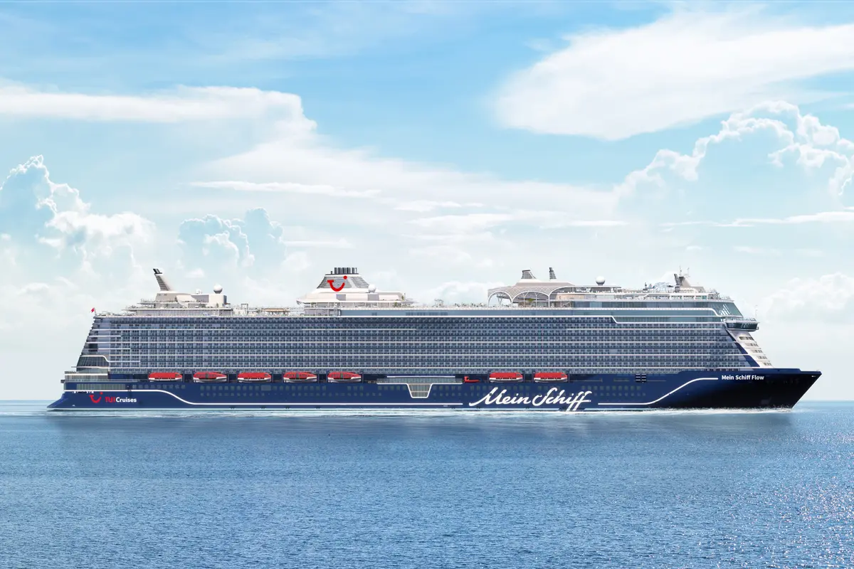 Seitenansicht der Mein Schiff Flow auf offenem Meer. Der blau-weiße Rumpf mit dem TUI Cruises Logo ist prominent, darüber die vielen Kabinenfenster und Decks. Wenige Wolken am hellblauen Himmel über dem Schiff.