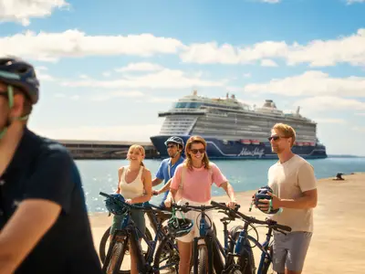Fünf lachende Personen stehen mit Fahrrädern und Helmen an einer Uferpromenade. Im Hintergrund ist die Mein Schiff Flow auf dem Meer zu sehen. Die Sonne scheint und der Himmel ist blau mit einigen Wolken.