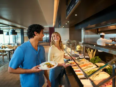 Zwei lächelnde Gäste am Buffet der Mein Schiff Flow. Ein Mann hält einen Teller mit Reis und Curry, eine Frau nimmt Salat. Im Hintergrund bereitet ein Koch Speisen zu.