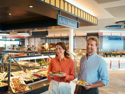 Ein glückliches Paar mit Tellern voller Essen vor einem großen Buffet im Restaurant an Bord der Mein Schiff Flow. Eine Frau in Orange und ein Mann in Hellblau lächeln. Das Buffet bietet eine reiche Auswahl an frischen Speisen und Salaten. Im Hintergrund sind weitere Gäste und das Restaurantambiente zu sehen.