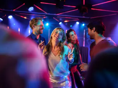 Vier junge Menschen tanzen und lachen bei einer Party an Bord der Mein Schiff Flow. Die Szene ist von farbigem Licht in Blau, Rot und Pink beleuchtet. Im Vordergrund sind schemenhaft weitere Personen zu sehen.