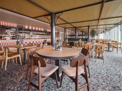 Innenansicht des Selbstbedienungsrestaurants an Bord der Mein Schiff Flow. Mehrere Buffets sind im Hintergrund sichtbar, wo Köche Essen zubereiten. Zahlreiche Tische und Stühle in hellbraunen Holztönen sind im Vordergrund und Mittelbereich verteilt. Der Boden ist mit einem bunten Teppich ausgelegt, und eine große Fensterfront erstreckt sich entlang der rechten Seite des Raumes.