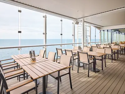 Sonniges Restaurant auf der Mein Schiff Flow mit Holzböden und zahlreichen Tischen und Stühlen. Die Panoramafenster bieten einen weiten Blick auf das offene Meer.