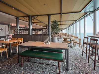 Ein weitläufiger Restaurantbereich an Bord der Mein Schiff Flow mit großen Fenstern zum Meer. Es gibt Holztische mit Stühlen und Bänken. Im Hintergrund sind weitere Tische und eine offene Küche sichtbar.