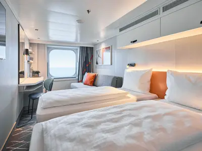 Eine helle Kabine an Bord der Mein Schiff Flow mit zwei Einzelbetten im Vordergrund, einem Sofa mit orangefarbenen Kissen, einem Schreibtisch und einem runden Fenster mit Meerblick.