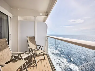 Sonniger Blick von einer Balkonkabine der Mein Schiff Flow auf das glitzernde blaue Meer und einen bewölkten Himmel. Zwei Stühle stehen auf dem Holzboden des Balkons.