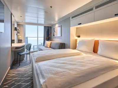 Eine helle und moderne Balkonkabine an Bord der Mein Schiff Flow mit einem Doppelbett, einem Sofa, einem Schreibtischbereich und einem Balkon mit Meerblick. Der Raum ist in neutralen Farben gehalten und wird durch Kunstwerke und indirekte Beleuchtung akzentuiert.