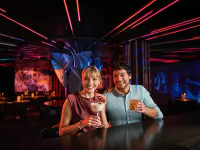 Ein lachendes Paar sitzt an einer Bar auf der Mein Schiff Flow. Die Frau hält einen cremefarbenen Cocktail, der Mann ein braunes Getränk. Im Hintergrund sind weitere Tische und bunte Lichtinstallationen zu sehen.