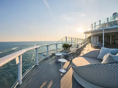 Blick über das Deck der Mein Schiff Flow bei Sonnenuntergang. Im Vordergrund sind bequeme Lounge-Möbel zu sehen. Weiter hinten reihen sich Liegestühle entlang der Reling mit Glasbrüstung. Die Sonne geht hell am Horizont unter und spiegelt sich auf dem Meer. Im Hintergrund ist ein Teil des oberen Decks mit einer Bar und weiteren Sitzgelegenheiten zu erkennen.