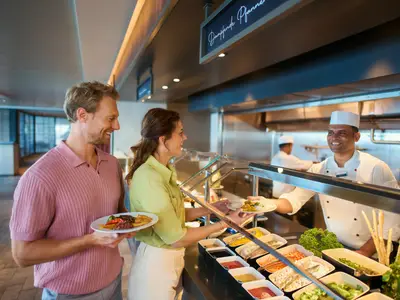 Zwei Gäste lächeln, während ein Koch ihnen Speisen an einem Buffet an Bord der Mein Schiff Flow reicht. Der Mann hält einen Teller, die Frau nimmt einen Teller vom Koch entgegen. Im Hintergrund ist die Schiffsküche zu sehen.