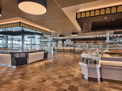 Ein weitläufiges, modernes Buffetrestaurant an Bord der Mein Schiff Flow mit mehreren Essensstationen, hellen Fliesenböden und großen Fenstern im Hintergrund.