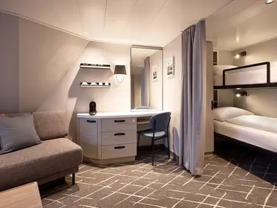 Innenansicht einer Kabine an Bord der Mein Schiff Flow, die ein Sofa mit Kissen, einen Schreibtisch mit Stuhl und einen abgetrennten Bereich mit einem Etagenbett zeigt.