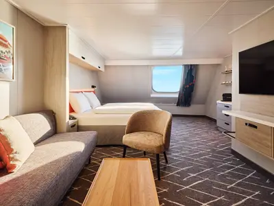 Eine helle Kabine der Mein Schiff Flow mit Doppelbett, Sofa, braunem Sessel, Holztisch und Fernseher. Ein Fenster zeigt blauen Himmel.