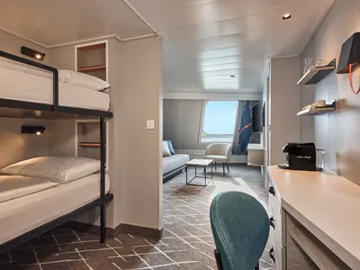 Innenansicht einer geräumigen Kabine der Mein Schiff Flow. Links sind zwei Kojenbetten mit weißen Bezügen zu sehen. Rechts führt ein Durchgang zu einem Wohnbereich mit Sofa, Sessel und einem Fenster mit Meerblick. Im Vordergrund rechts steht eine Kaffeemaschine auf einem hellen Sideboard. Die Wände sind in hellem Grau gehalten, der Teppichboden hat ein geometrisches Muster.