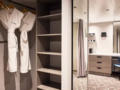 Detailansicht einer Kabine auf der Mein Schiff Flow. Links hängen zwei weiße Bademäntel. Rechts sind offene Regale zu sehen, daneben ein Waschtisch mit Schubladen, Nespresso-Maschine, Tassen und einem Spiegel. Ein blauer Stuhl steht davor.