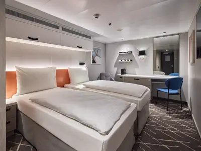 Ein helles Doppelzimmer an Bord der Mein Schiff Flow, das zwei getrennte Einzelbetten mit weißer Bettwäsche zeigt. Im Hintergrund befinden sich ein Schreibtisch mit Spiegel und ein blauer Stuhl.