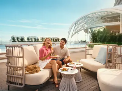 Ein junges, lachendes Paar sitzt an Bord der Mein Schiff Flow auf bequemen Outdoor-Möbeln und isst Brunch. Im Hintergrund sind ein Pool und eine moderne architektonische Struktur zu sehen. Der Himmel ist blau.
