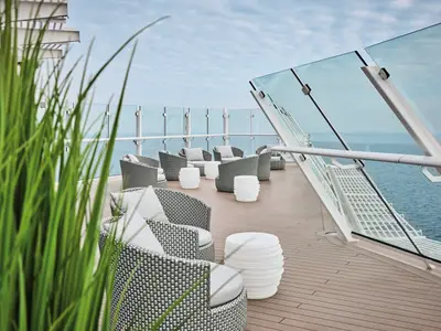 Blick auf das offene Deck der Mein Schiff Flow mit mehreren grauen Korbsesseln und runden weißen Tischen, umgeben von einer Glasbrüstung und mit grünem Gras im Vordergrund. Das Meer ist im Hintergrund sichtbar.
