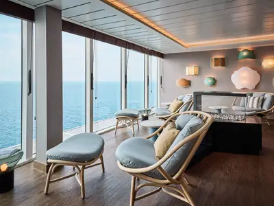 Sonnendurchflutete Lounge auf der Mein Schiff Flow mit großen Fenstern zum Meer. Mehrere moderne Sessel und Hocker sind im Raum verteilt, während eine Wand mit verschiedenen Leuchten in kunstvollen Formen dekoriert ist.