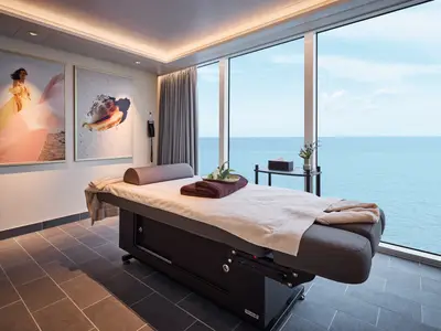 Ein luxuriöser Spa-Behandlungsraum an Bord der Mein Schiff Flow. Eine Massagebank mit Handtüchern und einer Schale mit Blumen steht vor einem raumhohen Fenster mit Blick auf das weite, blaue Meer. An der Wand hängen zwei große Bilder: eines einer Frau am Strand und eines einer Muschel. Die Beleuchtung ist warm und einladend.