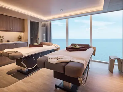 Ein luxuriöser Spa-Raum an Bord der Mein Schiff Flow mit zwei Massageliegen, bereit für Behandlungen. Durch die großen Fenster ist eine weite Meereslandschaft zu sehen. Eine Duschkabine ist im Hintergrund links sichtbar.